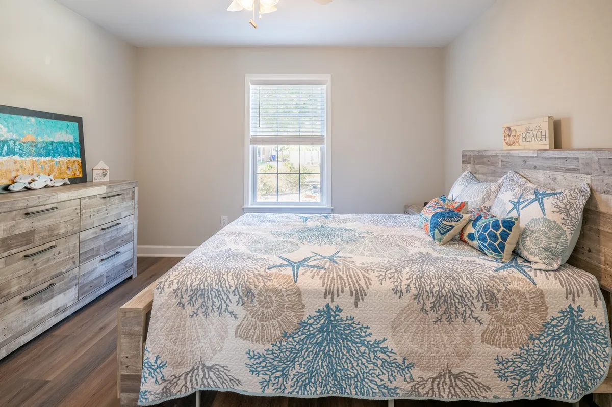 A serene guest bedroom with a bed dressed in a coral-patterned quilt, a wooden dresser, and abundant natural light streaming through the window. The design is simple and welcoming.