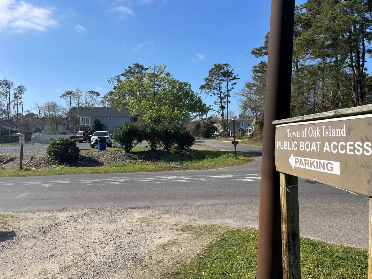 This image captures a public boat access sign, several parked vehicles, and a backdrop of trees and a roadway. The setting highlights easy access to water activities such as boating and fishing, with parking for cars and trailers visible.