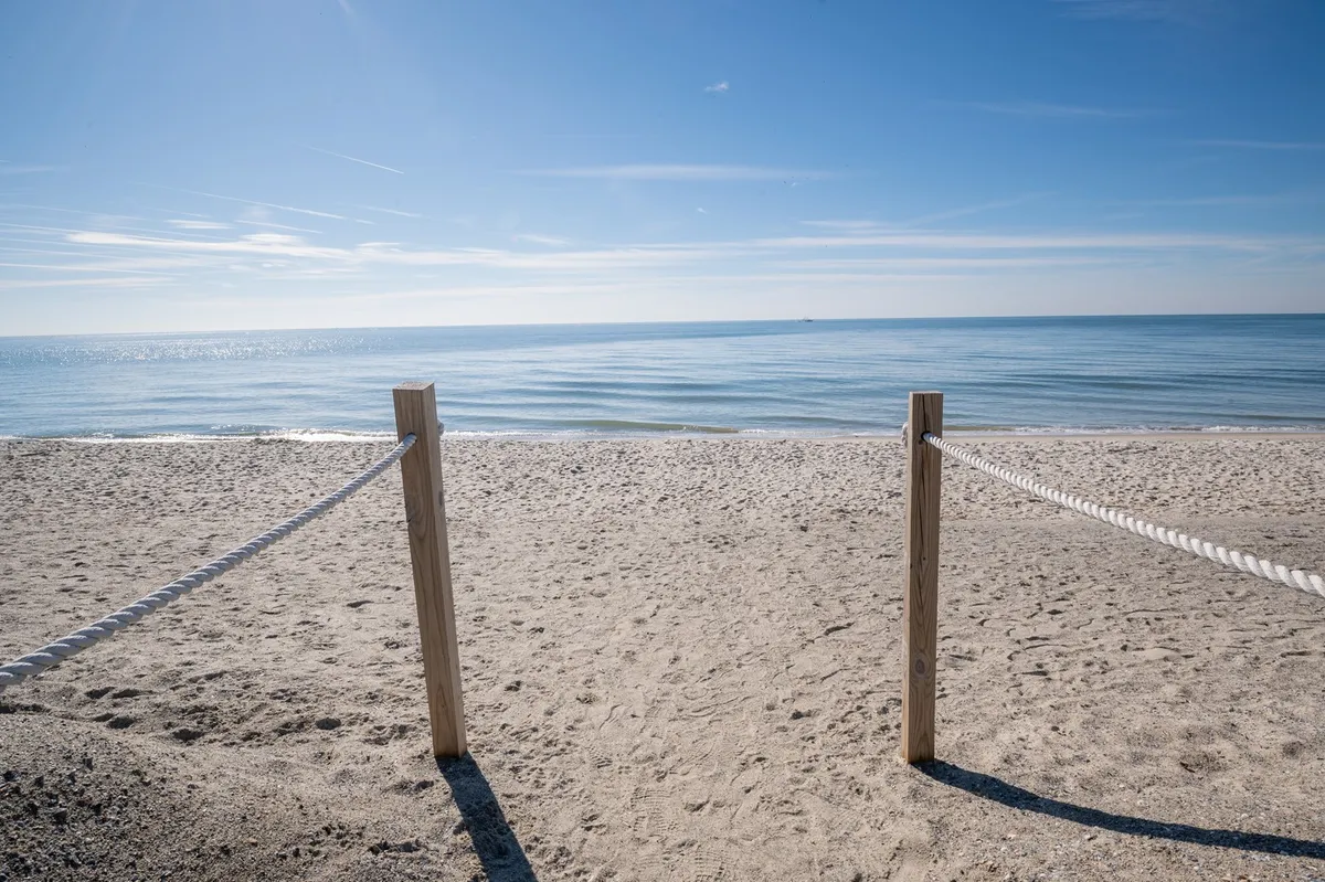 A tranquil sandy beach with wooden posts and a rope barrier, calm blue sea in the background, and plenty of open space for families to relax, sunbathe, or play. The shoreline is inviting and ideal for family activities like swimming or building sandcastles.