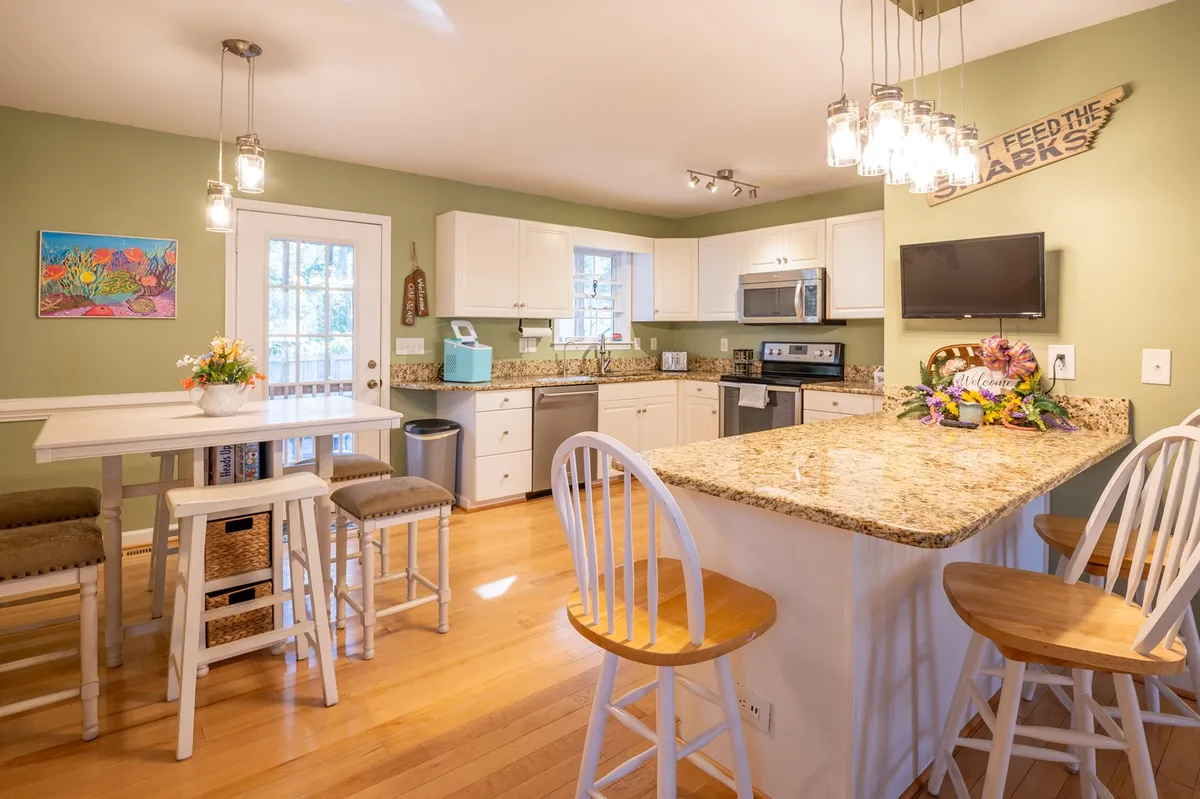 The image displays a bright kitchen with white cabinetry, a granite countertop, kitchen island with bar stools, and colorful artwork. The seating at the island highlights the social aspect of the kitchen, inviting interaction and casual dining.