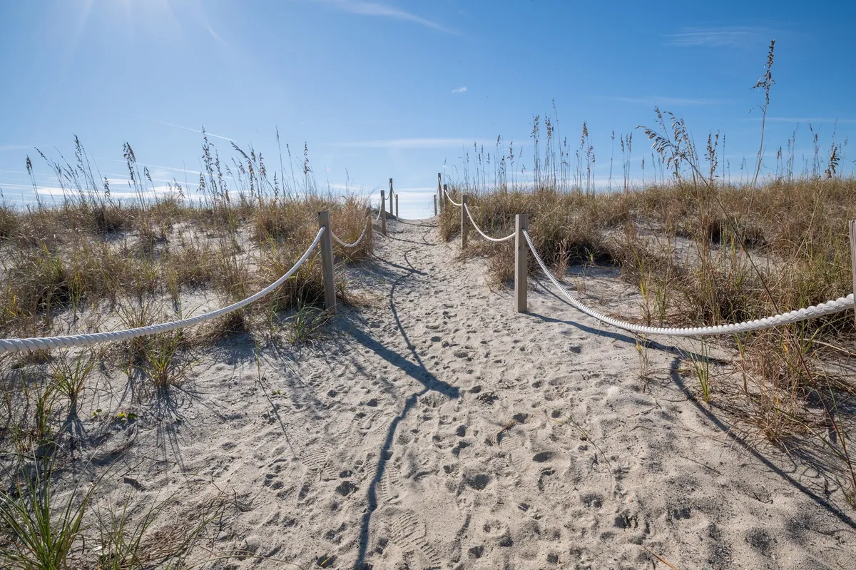 A sandy pathway lined with tall grasses and rope fencing leads toward the ocean, evoking the anticipation of a leisurely walk to the beach. The tranquil scene invites viewers to imagine the short, scenic stroll from the cottage to the shore.