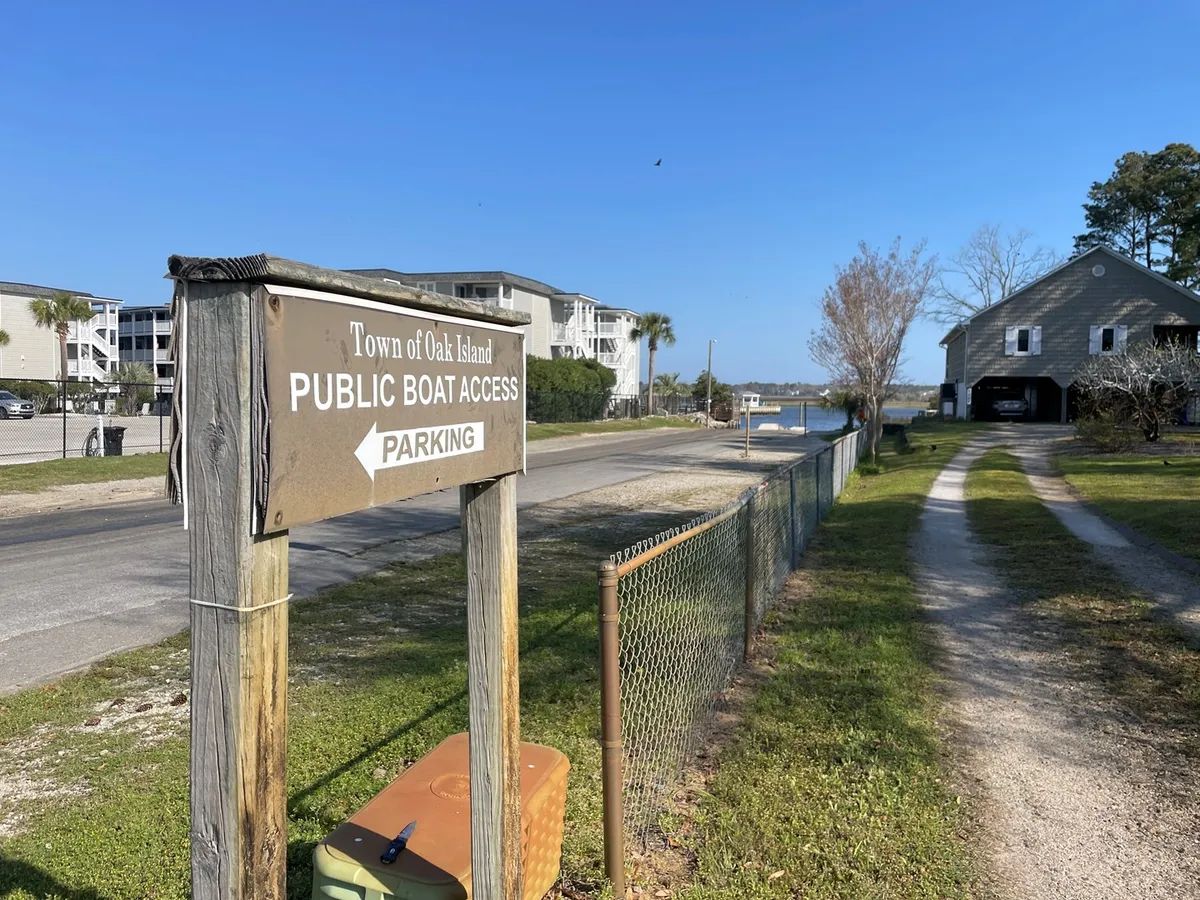 An inviting scene showing a public boat access sign and a pathway leading toward the water, surrounded by nearby buildings and a parking area. The gentle slope of the path opens up to a tranquil shore, perfect for picnicking, fishing, or setting off on a boating adventure. The surroundings evoke the casual, community-focused lifestyle of Oak Island and hint at the area's local recreation.
