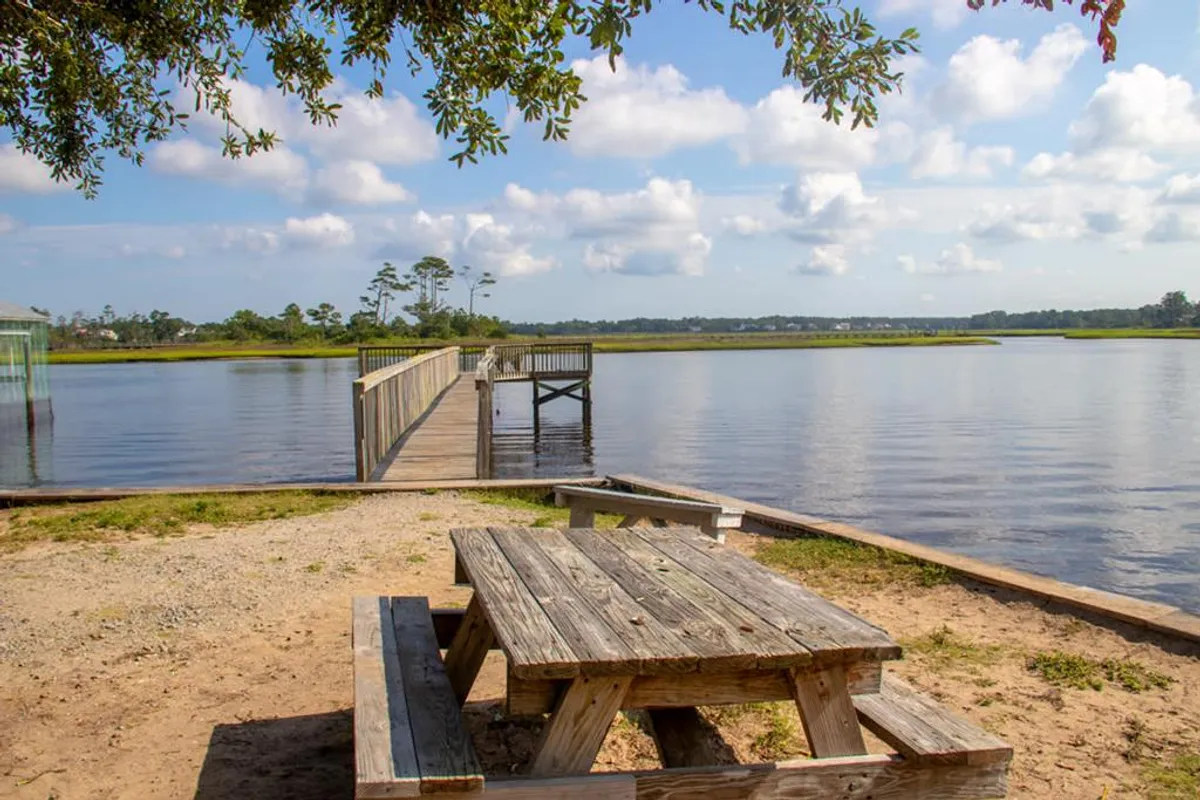 A tranquil exterior scene featuring a picnic table by the water's edge, a wooden dock, and serene water views. The setting is ideal for relaxed picnics, fishing, or simply enjoying the peaceful outdoors, tying the home to its scenic, accessible surroundings.