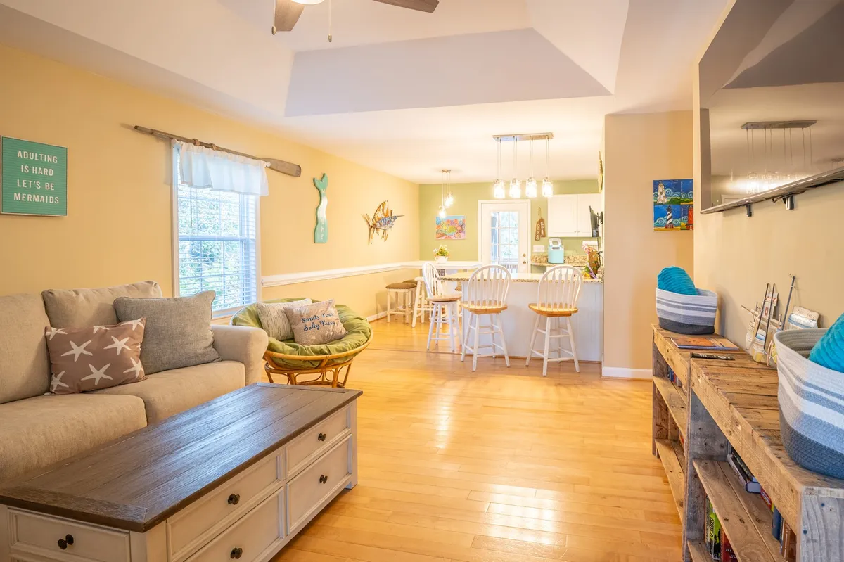 A welcoming open-concept space blending the living room and kitchen bar. Features a sofa accented with decorative pillows, a wooden coffee table, bar stools at the kitchen counter, and eye-catching wall art. The layout encourages seamless movement and sociability.