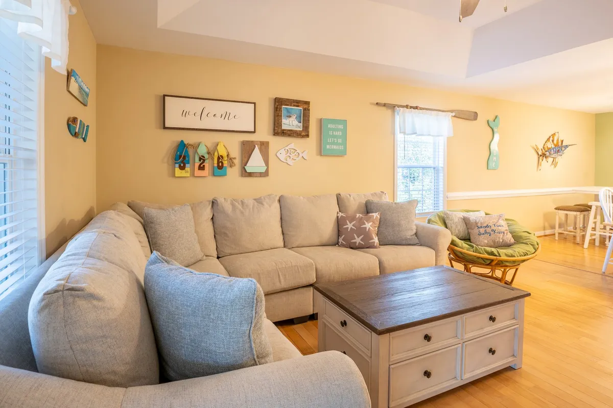 A spacious, open-concept living room featuring a large sectional sofa with soft cushions, a stylish coffee table, decorative wall art, and a green accent chair. The room is softly lit and invites relaxation, perfect for gatherings and cozy family moments.