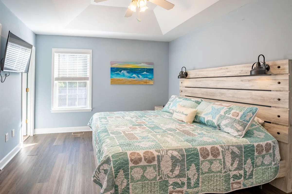 A cozy bedroom featuring a bed with a patterned quilt, artwork on the wall, and a TV mounted for entertainment. Sunlight filters softly through a nearby window, creating a peaceful atmosphere ideal for relaxation.