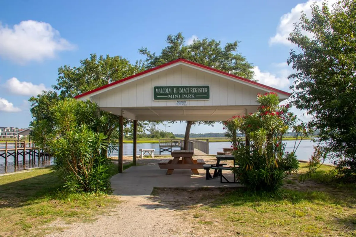 A cheerful pavilion sits amidst lush greenery with picnic tables positioned to overlook a sparkling water view. The sign indicates a welcoming communal space, and the area is perfect for sunset gatherings, family celebrations, or taking part in community events. The relaxed, open setting embodies the spirit of local culture and year-round events.